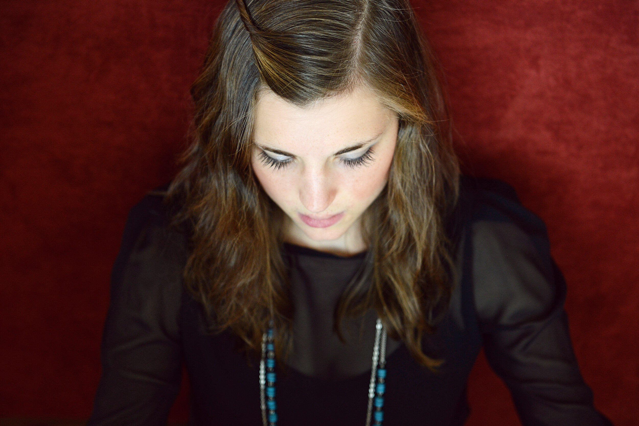 photo by Nir Arieli, image description: Headshot of Molly, a white female with brown hair, taken from above. She is looking forward and wearing a black shirt with a blue necklace, and is against a red background.