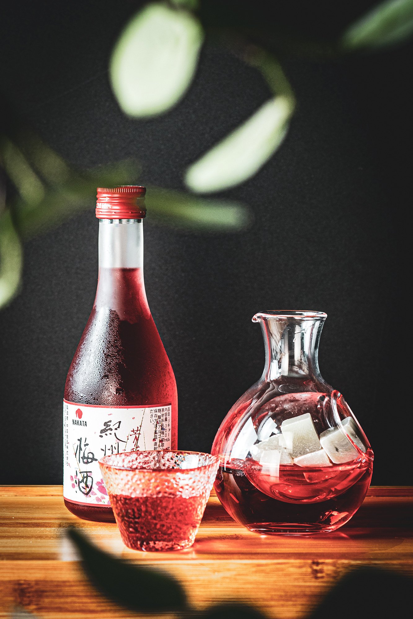 Bottle, glass, and decanter with red liquid and ice cubes on a wooden surface.